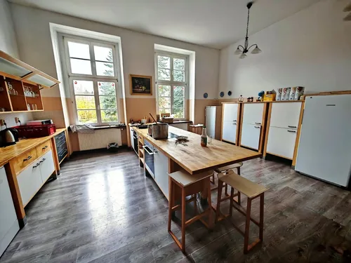 Großküche mit langem Arbeitstisch, mehreren Kühlschränken und großen Fenstern